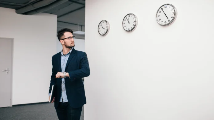 young-guy-glasses-is-walking-office-he-wears-blue-shirt-dark-jacket-jeans-beard-he-holds-laptop-hand-he-is-looking-clock-wall